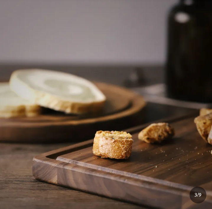 A wooden cutting board with a dark wood grain finish, shown in a styled kitchen setting with sliced white cheese.